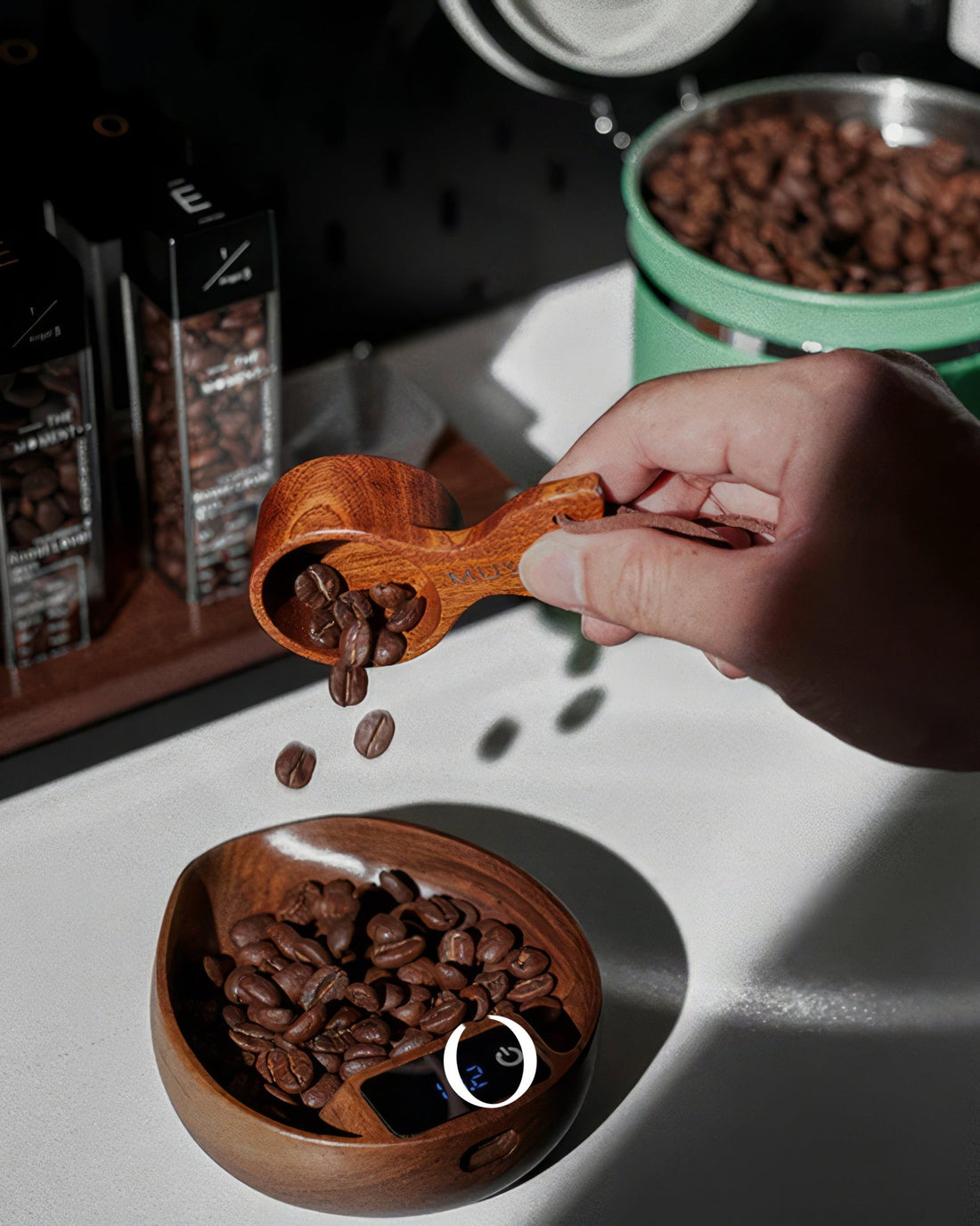 Hand pouring coffee beans from wooden scoop into wooden bowl with digital scale, surrounded by coffee containers and green tin of beans on white surface