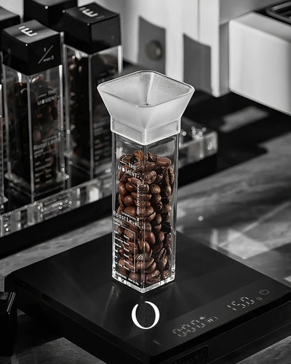 Clear glass container filled with coffee beans on a digital scale, surrounded by other coffee equipment, showcasing MUVNA coffee bean storage and display system