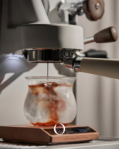 Espresso machine brewing coffee into glass cup on MUVNA walnut wood digital scale, showcasing precision and elegance in coffee preparation