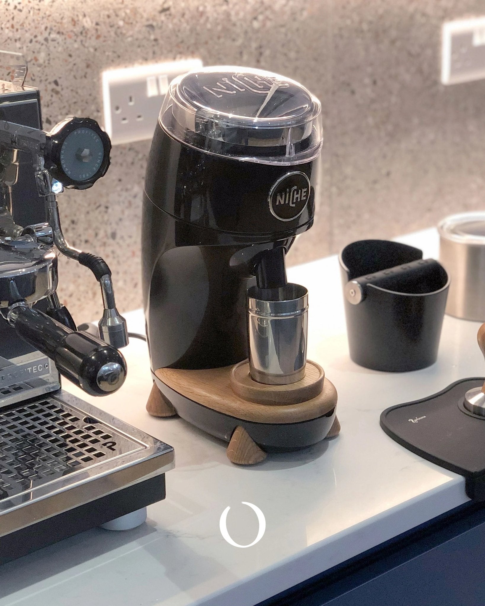 Niche Duo coffee grinder with sleek black design, positioned on wooden base next to espresso machine and accessories on countertop. Professional-grade home coffee setup featuring grinder, portafilter, and knock box.