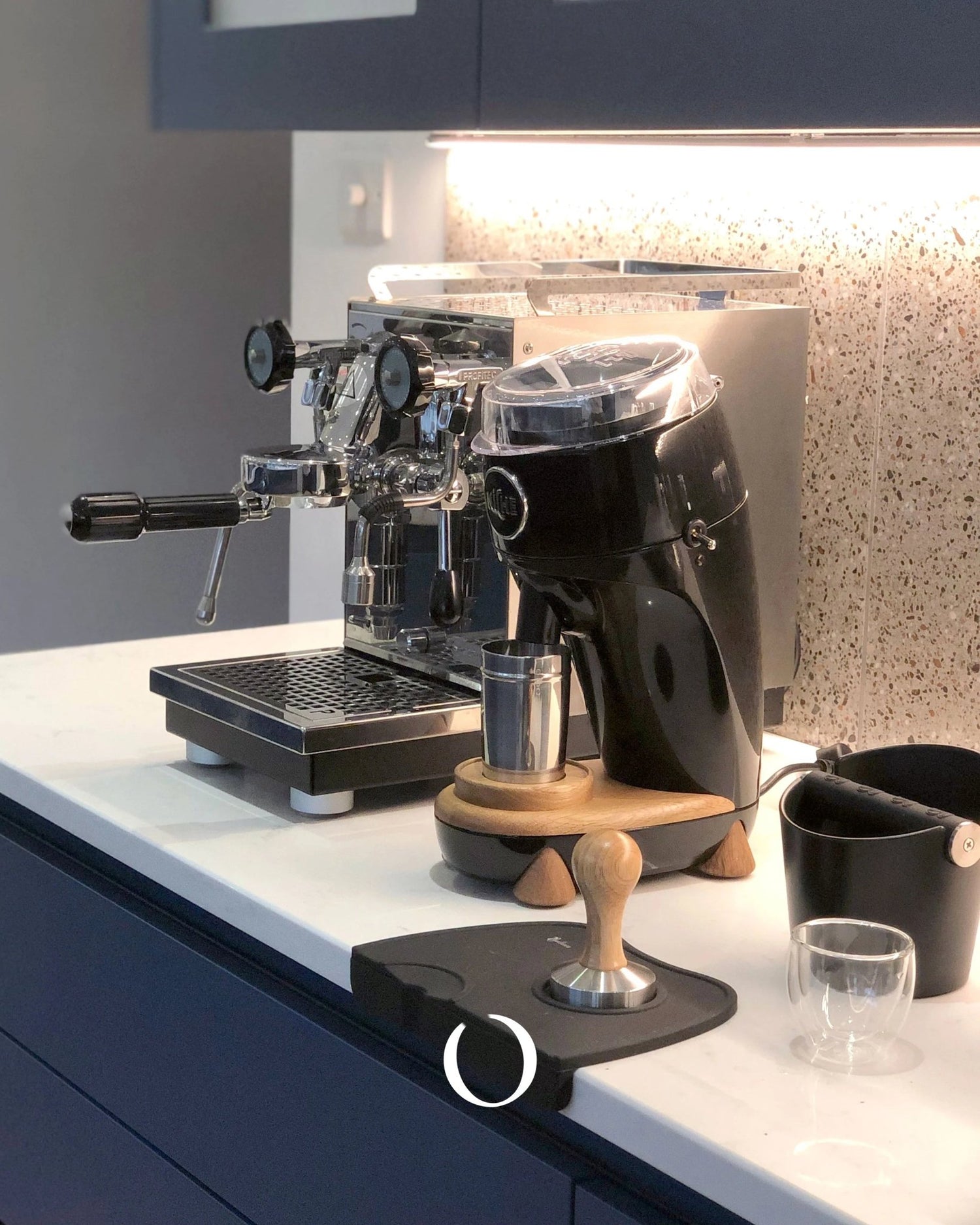 Sleek espresso machine and grinder setup on modern kitchen counter with under-cabinet lighting, tamper, and accessories for brewing premium coffee at home