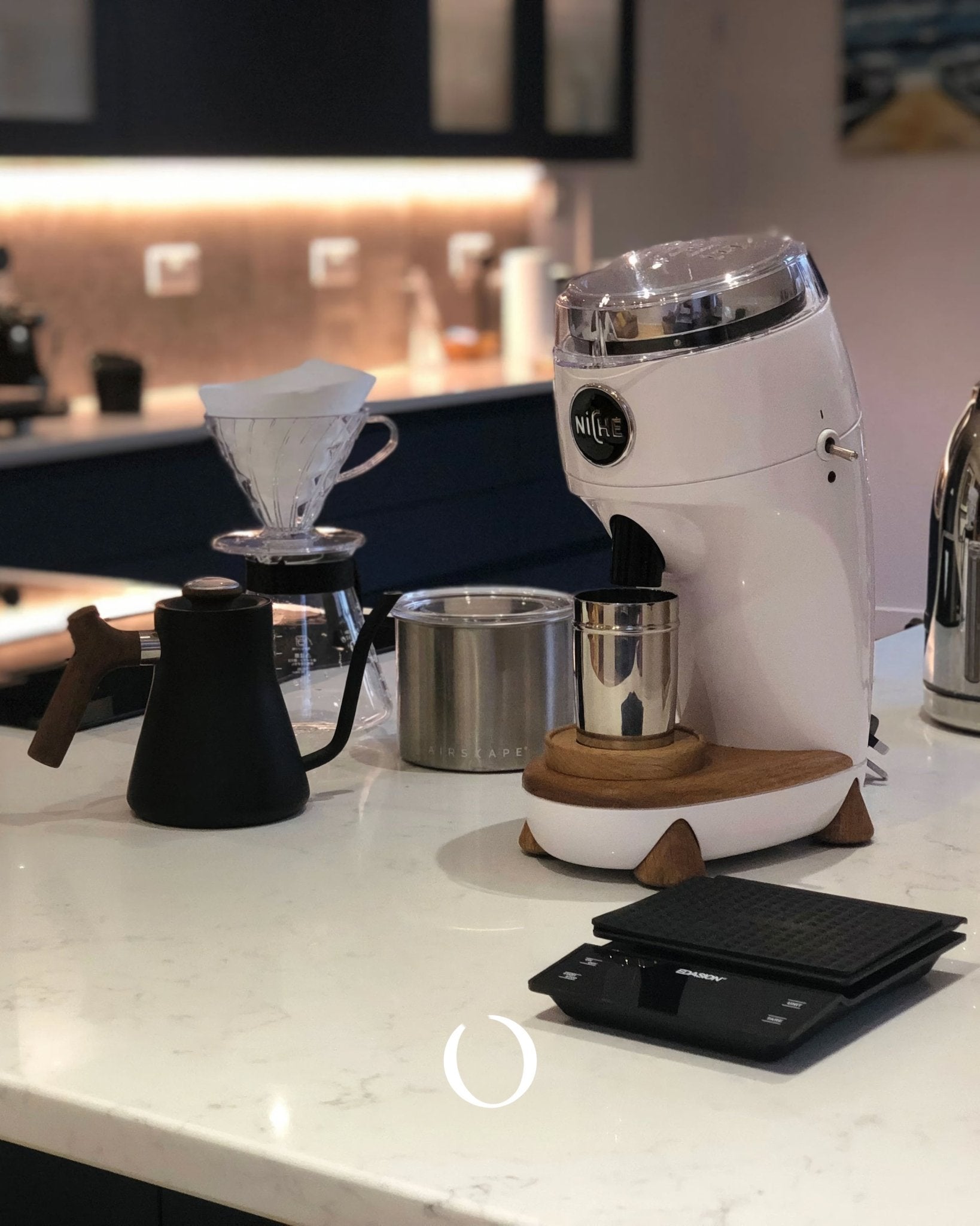 Niche Duo coffee grinder on kitchen counter with pour-over setup, stainless steel containers, and digital scale, showcasing professional home brewing equipment in modern kitchen setting