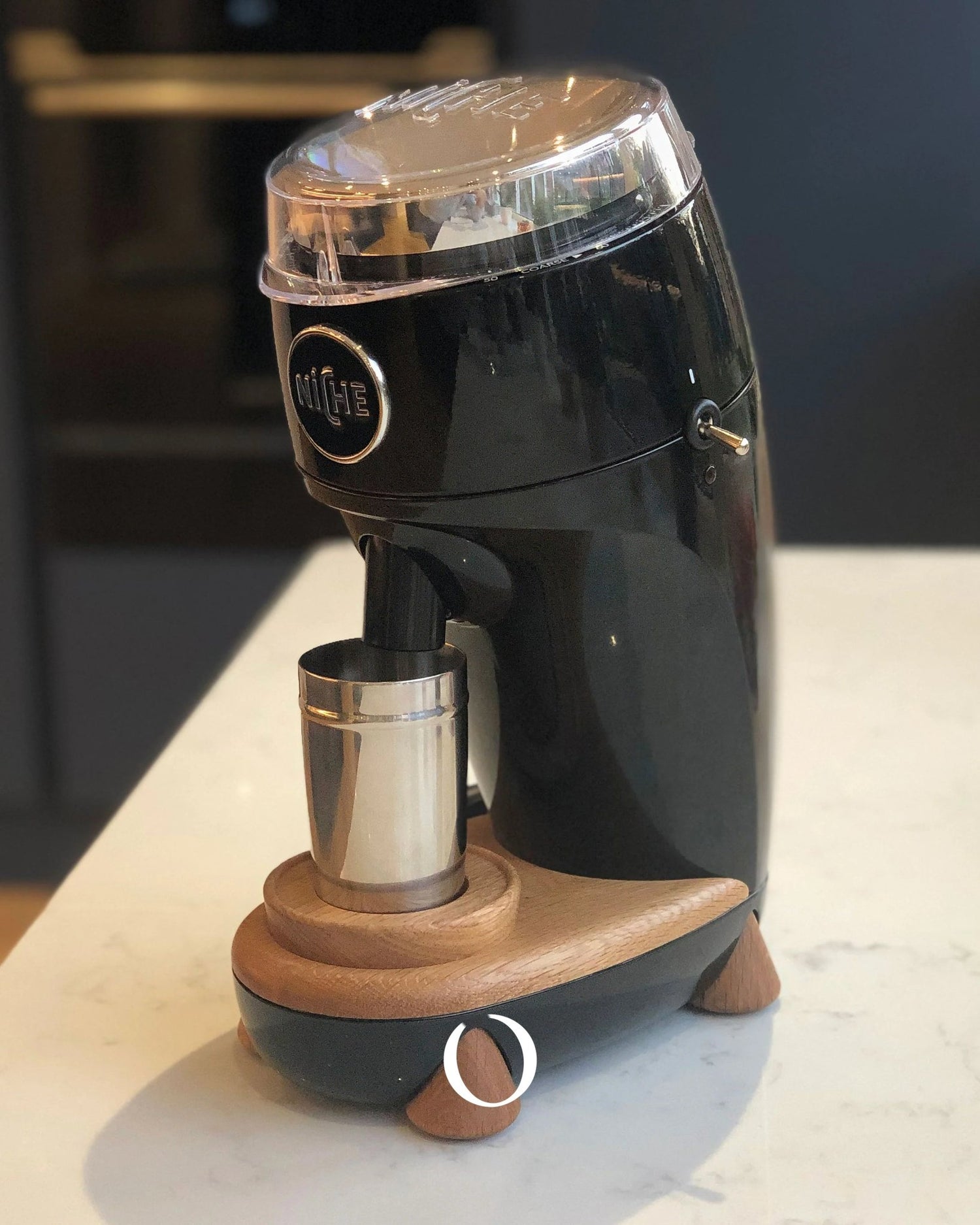 Niche Duo coffee grinder with sleek black body, transparent hopper, wooden base, and stainless steel grind cup on white countertop, showcasing professional-grade design for home use