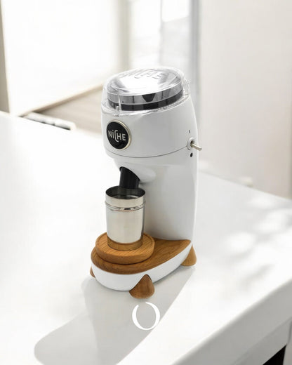 Niche Zero coffee grinder in white with wooden base, featuring sleek design and metal dosing cup, positioned on bright countertop with sunlight casting shadows