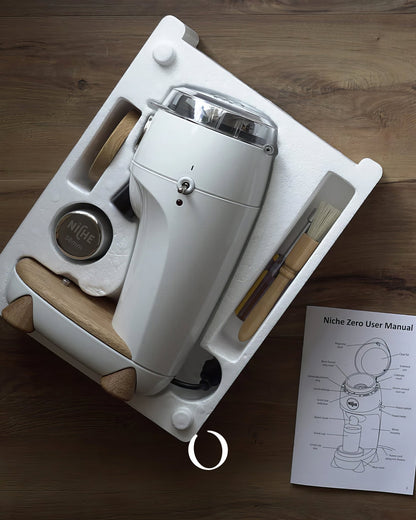 Niche Zero coffee grinder unboxed, showing white grinder, wooden accessories, user manual, and packaging on wooden surface