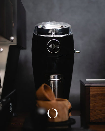 Sleek black Niche Zero coffee grinder with silver top and logo, positioned on counter with wooden accessories, showcasing modern design and professional-grade grinding capability for home baristas.