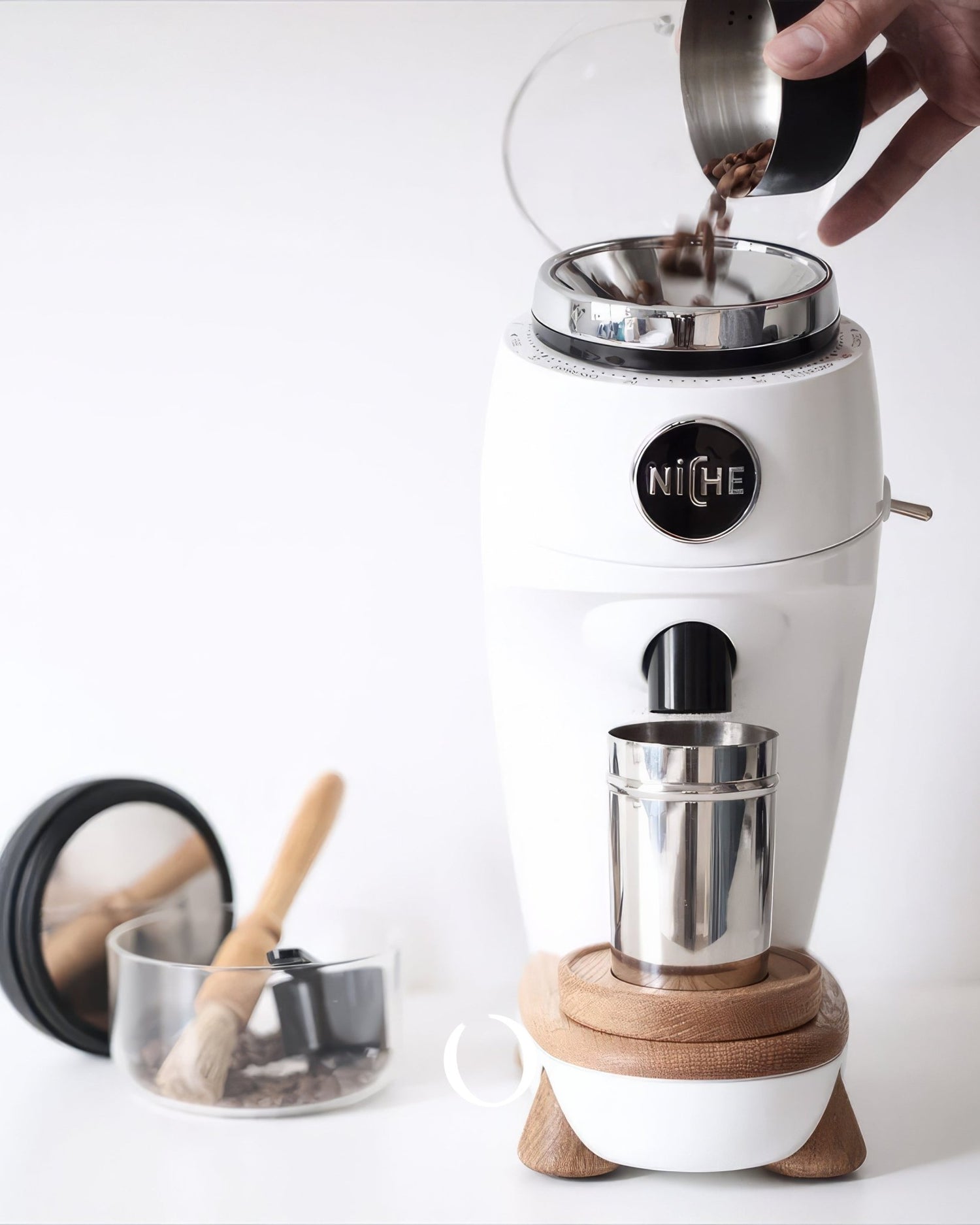 White Niche Zero coffee grinder with wooden base, pouring coffee beans into top chamber, stainless steel cup below, and coffee accessories in background