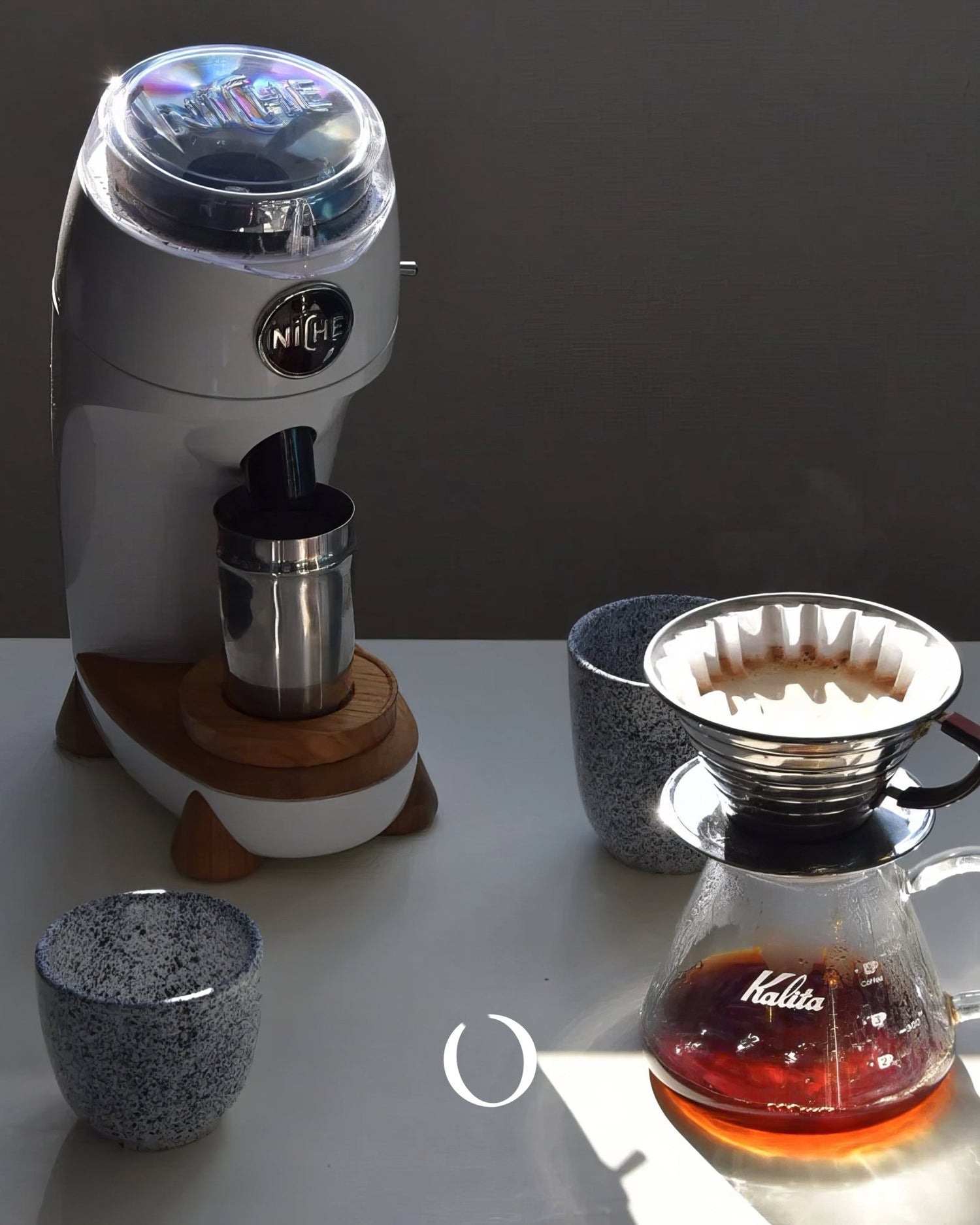 Niche Zero coffee grinder with wooden base, stainless steel cup, and pour-over coffee setup including Kalita dripper and carafe on a white surface