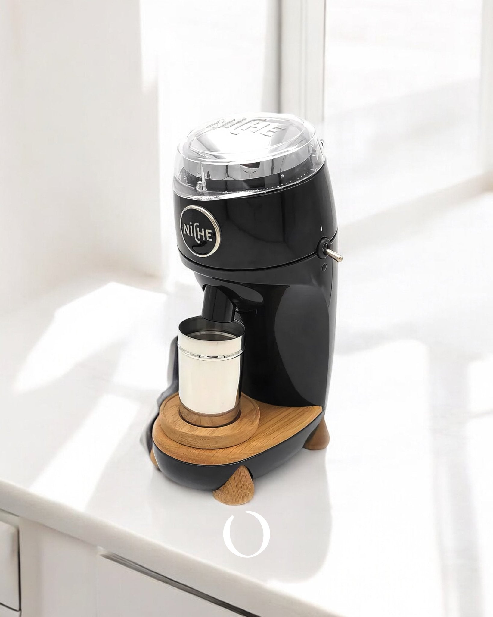 Niche Zero coffee grinder in black with wooden base, featuring a modern design and compact size, placed on a white countertop near a window, with a cup positioned underneath for freshly ground coffee.