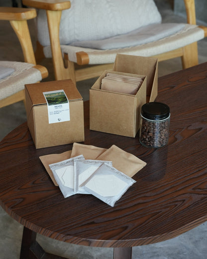 Coffee product packaging on wooden table including cardboard boxes, drip bags, and jar of coffee beans, with armchair in background