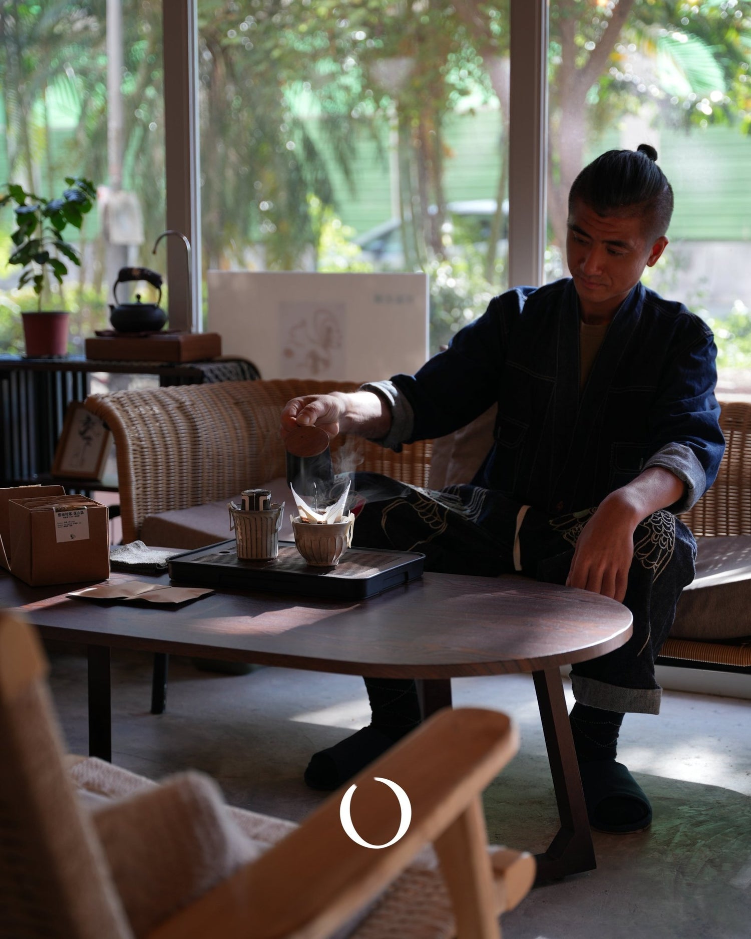 Person preparing pour-over coffee at a table in a sunlit room with plants and wicker furniture, demonstrating the artisanal brewing process of Santos Brazil coffee from Sip Coffee