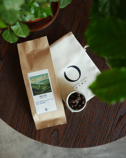 Brazilian coffee product display with two packages, one featuring a landscape image, alongside a small cup of coffee beans on a wooden surface, surrounded by green plant leaves