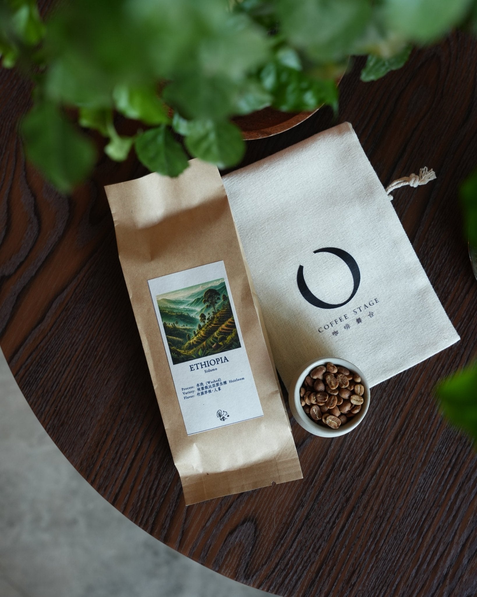 Coffee packaging from Sidamo, Ethiopia, featuring a brown paper bag with a landscape image, a white fabric pouch with a logo, and a small bowl of coffee beans on a wooden surface, with green plant leaves in the foreground.