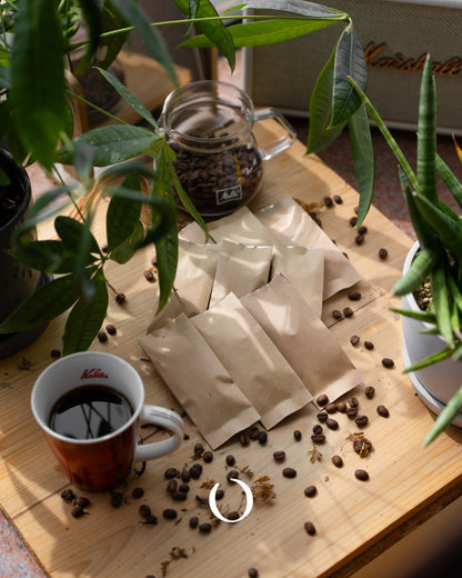 Coffee sampling setup with eight paper packets, coffee beans, a jar, and a red mug on a wooden board surrounded by green plants