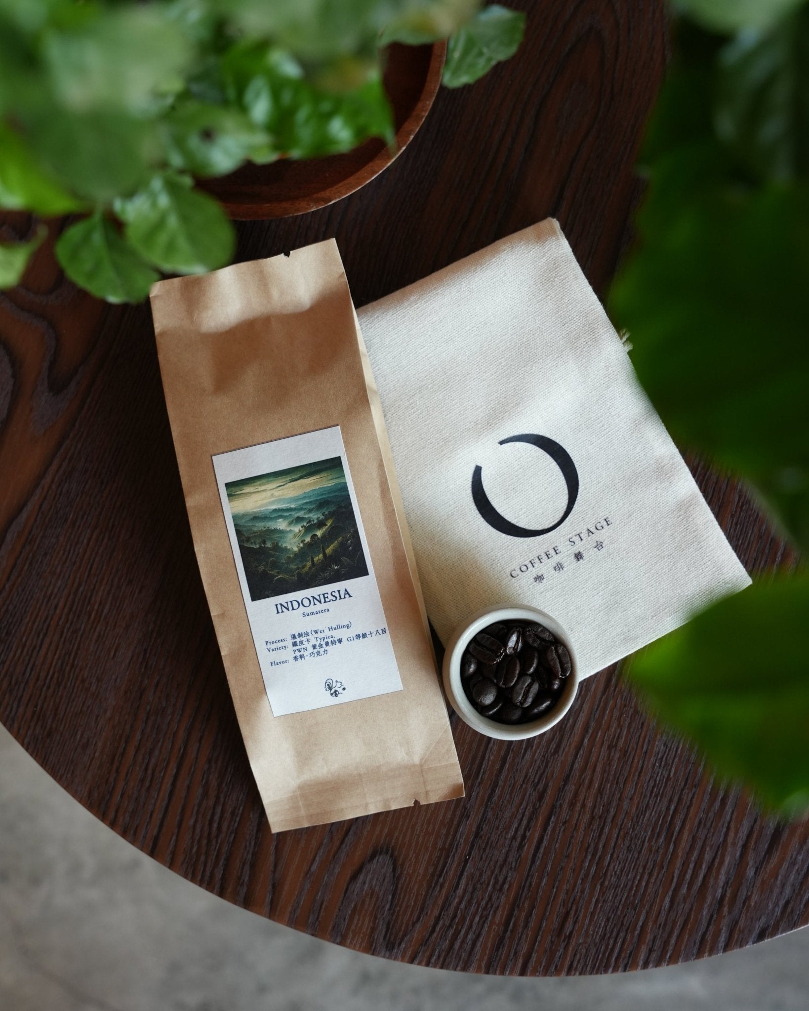 Artisanal Indonesian coffee presentation featuring a brown paper bag of Sumatera beans, a white branded pouch, and a small bowl of dark roasted coffee beans on a wooden surface with green plant leaves in the foreground.