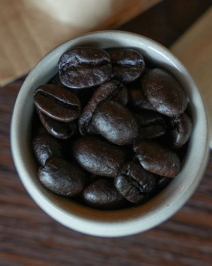 Ultra dark roast Sumatran coffee beans in a white ceramic cup, showcasing their rich, oily sheen and deep brown color, emphasizing the intense roast level of the Typica variety from Indonesia