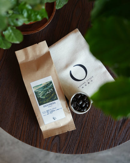 Two coffee bags, one labeled Indonesia with mountain image, next to small cup of coffee beans on wooden surface with green plant in foreground