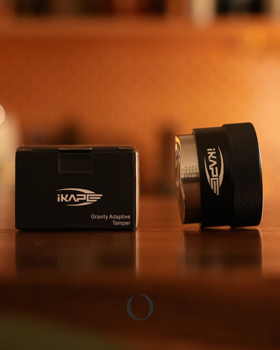 IKAPE Gravity Adaptive Tamper for espresso coffee, black metal device with packaging box on wooden surface, warm lighting atmosphere