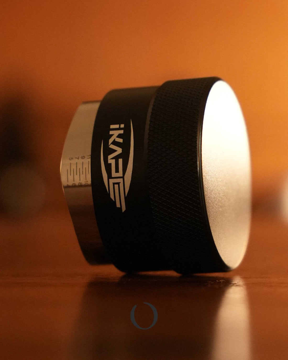 IKAPE espresso gravity distribution tool, black and silver cylindrical device for even coffee grounds distribution, shown on warm-toned surface in soft lighting