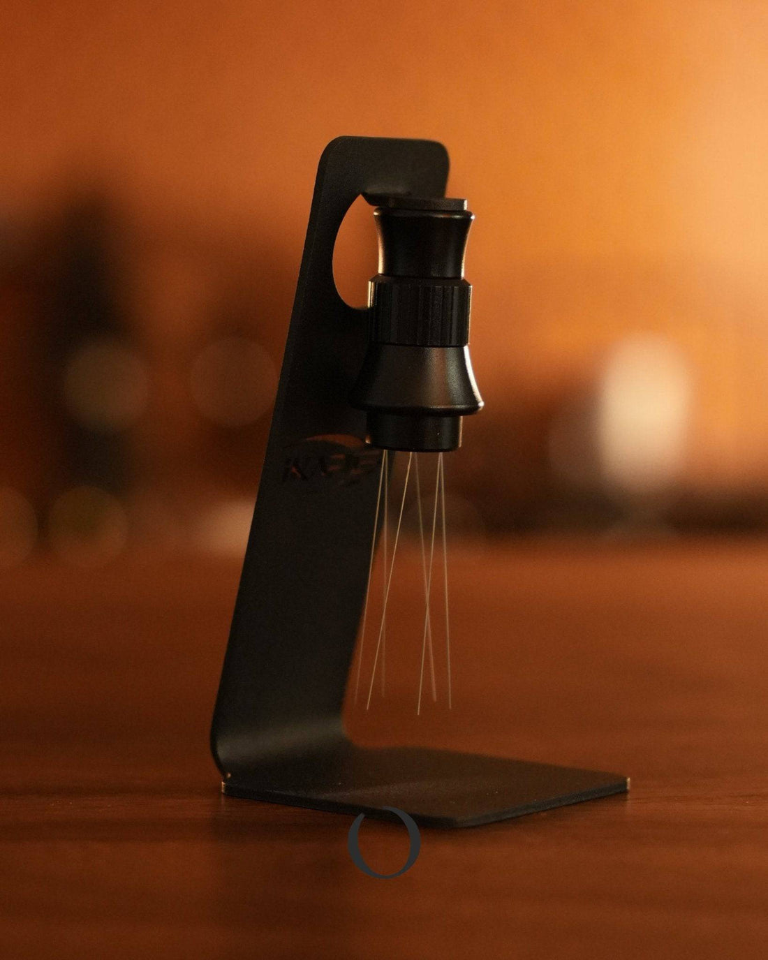 Ikape adjustable espresso distribution needle with magnetic design, showcasing its sleek black stand and innovative needle mechanism against a warm, orange background, highlighting its professional-grade functionality for precise coffee preparation.