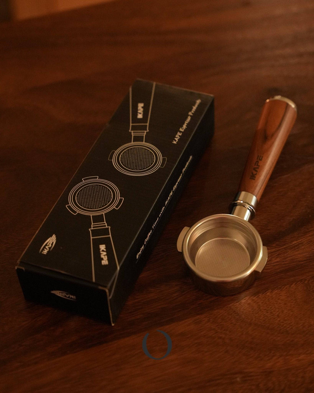 IKAPE 58mm bottomless portafilter with wooden handle and stainless steel filter basket, displayed next to its black product box on a wooden surface in warm lighting