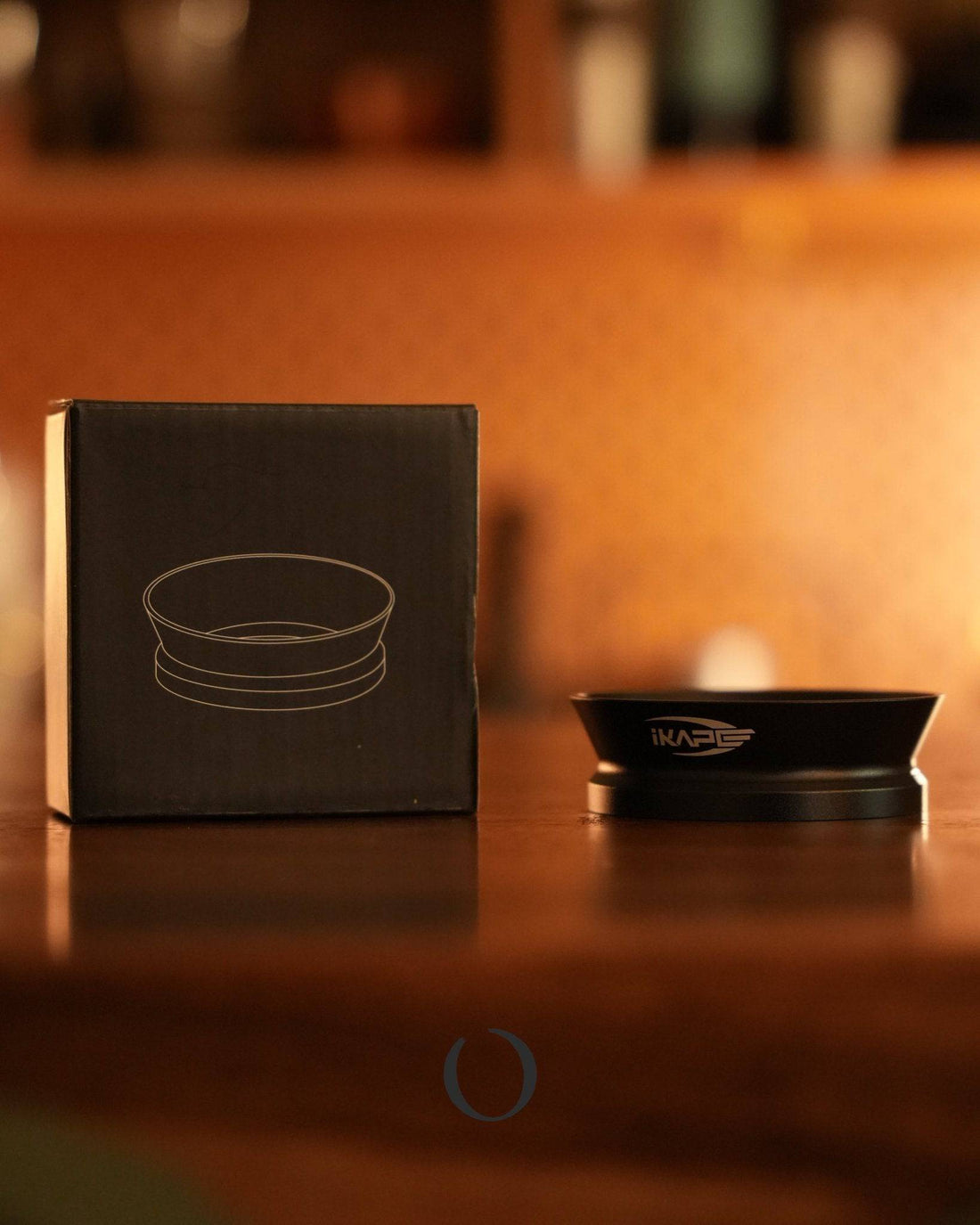 IKAPE V2 magnetic coffee dosing ring with its packaging box on wooden surface, showcasing sleek design and professional-grade quality for espresso preparation in warm, inviting cafe ambiance