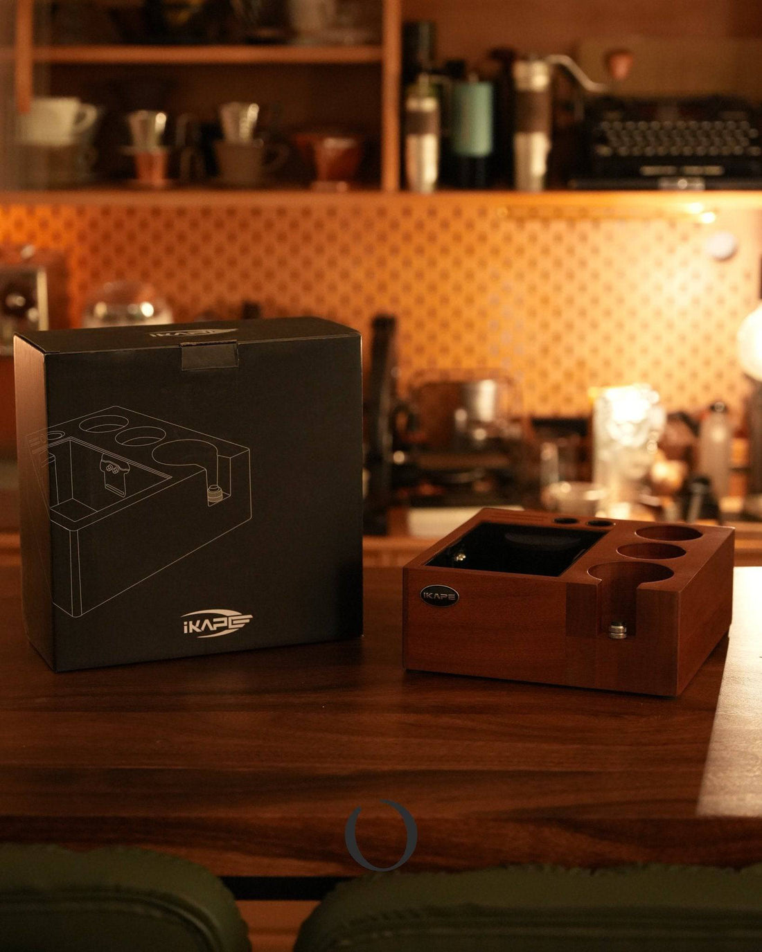 Ikape V2 Espresso Knock box with packaging on wooden counter in cozy coffee corner, showcasing elegant walnut design, multiple compartments for barista tools, and sleek black box with product illustration