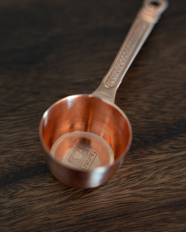 Kalita copper coffee scoop with laurel wreath pattern, 10g capacity, elegant rose gold finish, resting on dark wooden surface, showcasing precise measurement and artistic design for coffee enthusiasts