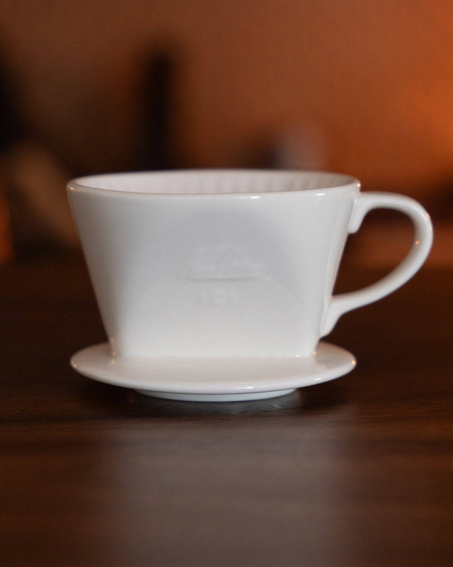 Kalita ceramic pour-over coffee dripper 101 in white, featuring a conical shape and handle, placed on a wooden surface with warm lighting, showcasing its elegant design for brewing specialty coffee