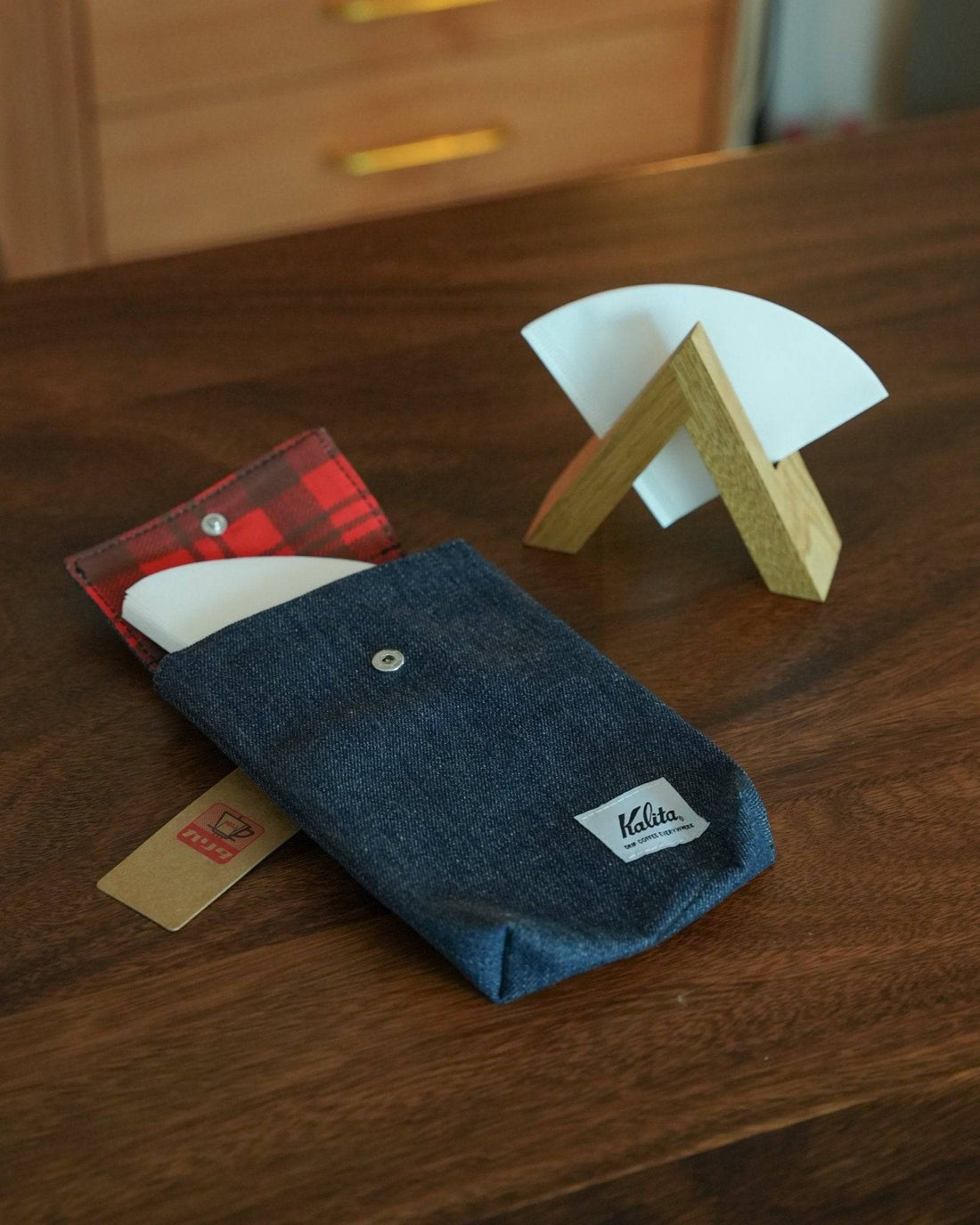 Kalita coffee filter storage bag in denim blue with red plaid lining, displayed next to wooden filter holder and white paper filter on wooden table