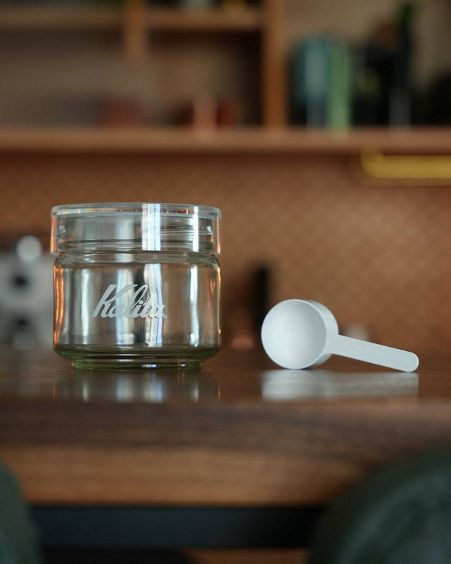Kalita glass coffee storage jar with airtight lid and white measuring scoop on wooden surface, kitchen background