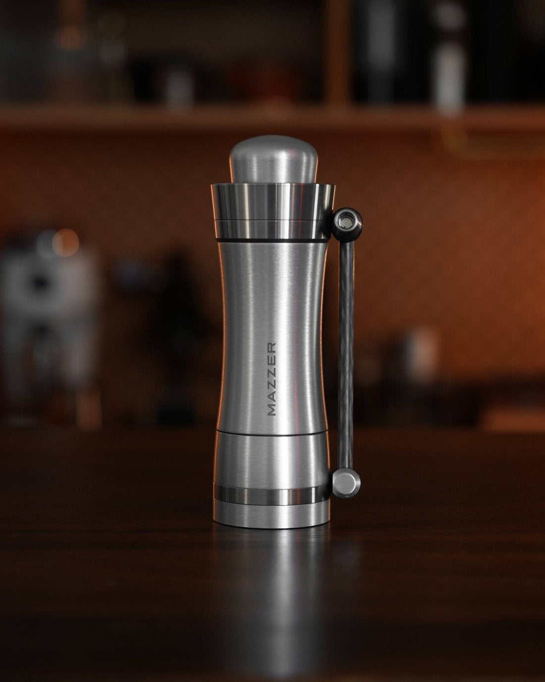 Mazzer Omega Fast manual coffee grinder, sleek stainless steel design with carbon fiber handle, standing on wooden counter in kitchen setting