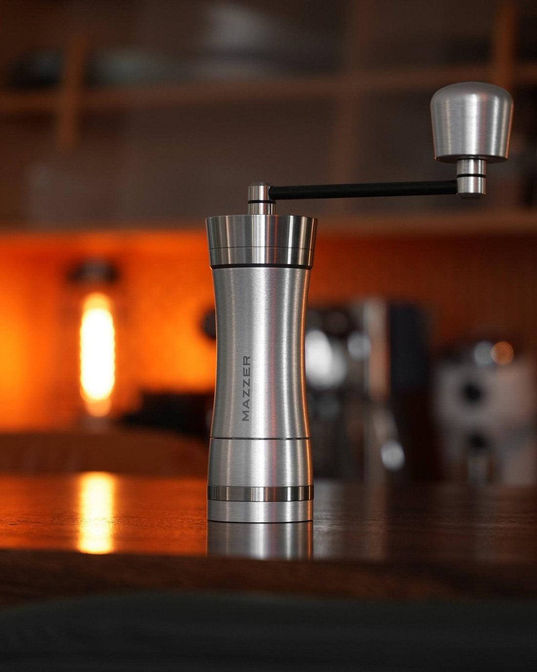 Mazzer Omega Soft manual coffee grinder on wooden table with warm ambient lighting, showcasing sleek stainless steel design and ergonomic handle for precision grinding