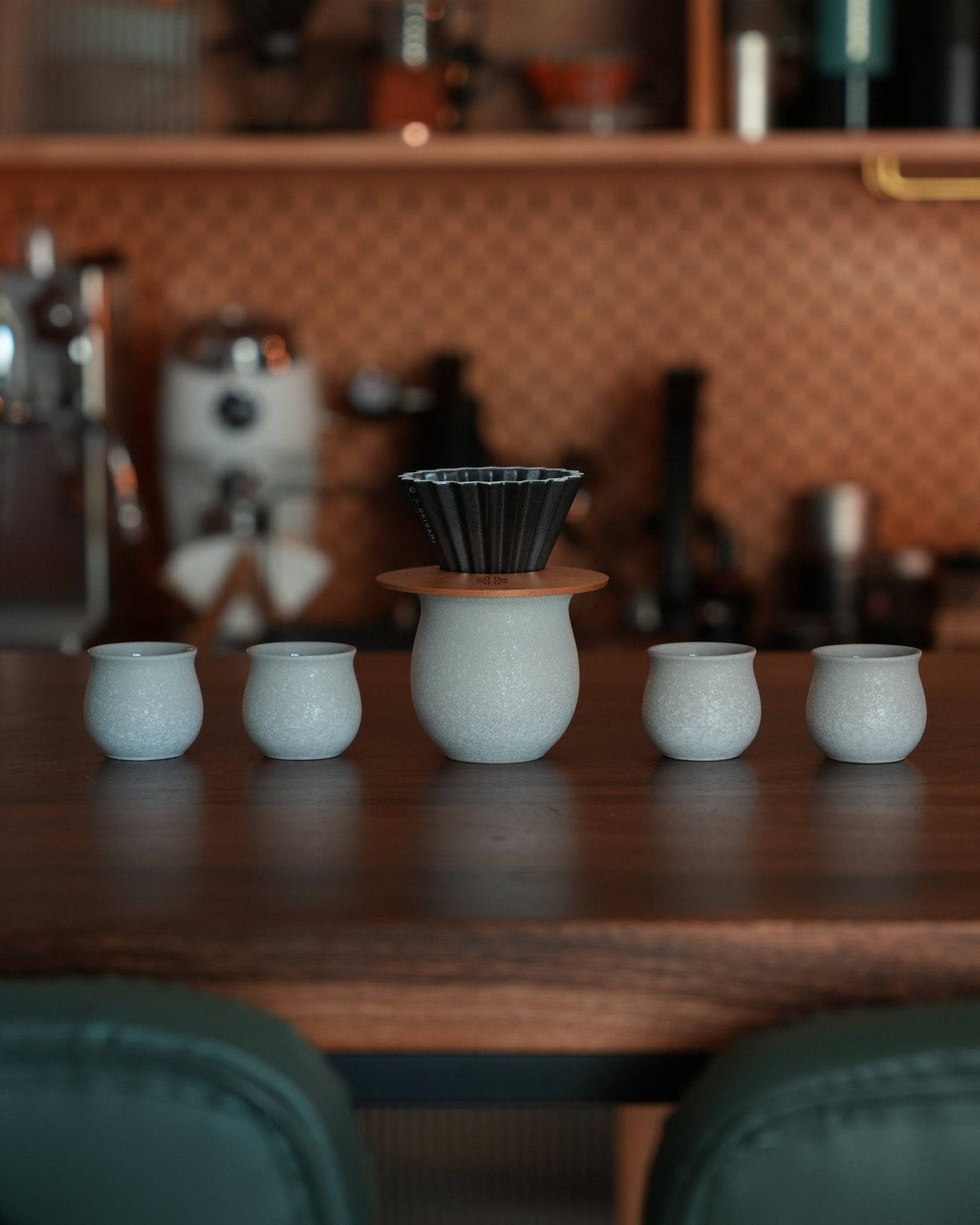 Ninety Plus coffee brewing set featuring a black pour-over dripper on a white ceramic base, surrounded by four matching white ceramic cups, displayed on a wooden counter in a cozy coffee shop setting