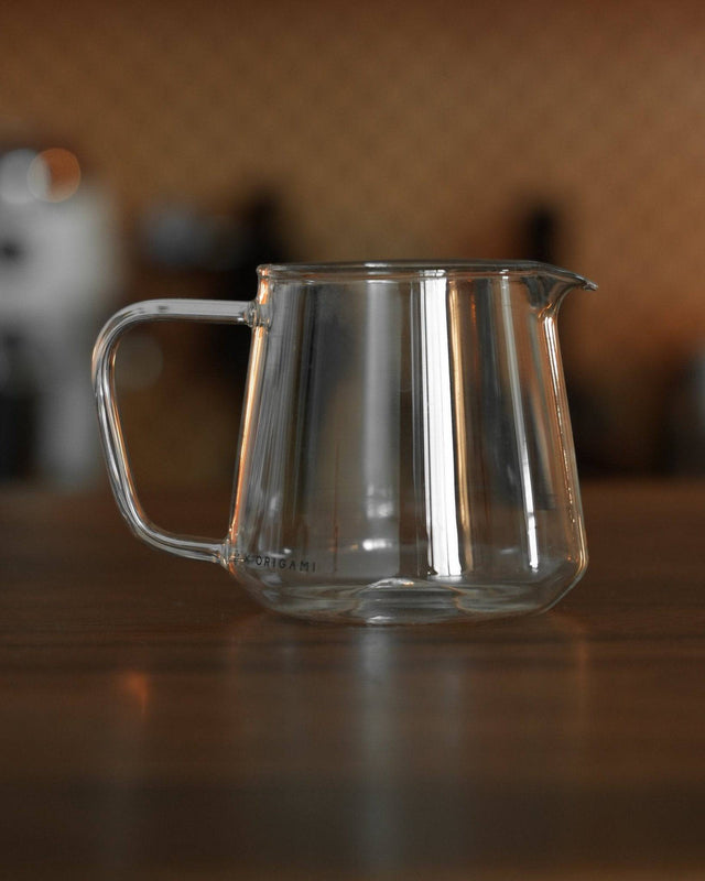 Clear glass Ninety Plus Coffee serving pitcher on wooden surface, showcasing sleek design with wide spout and ergonomic handle, perfect for precise pouring of premium coffee