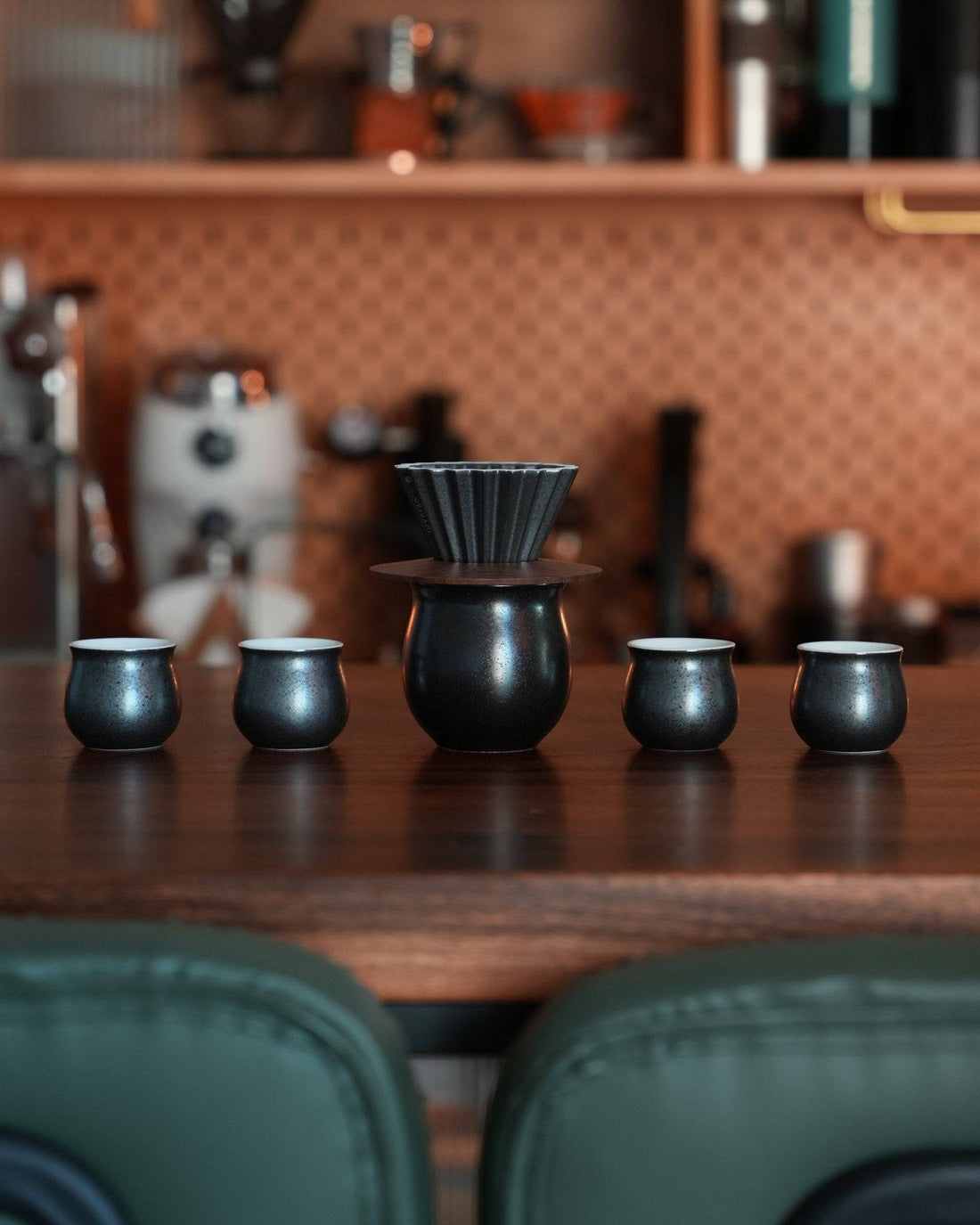 Ninety Plus pour-over coffee set in obsidian black, featuring a ceramic dripper and four matching cups, displayed on a wooden counter in a stylish coffee shop setting with copper-toned backsplash and green chairs visible