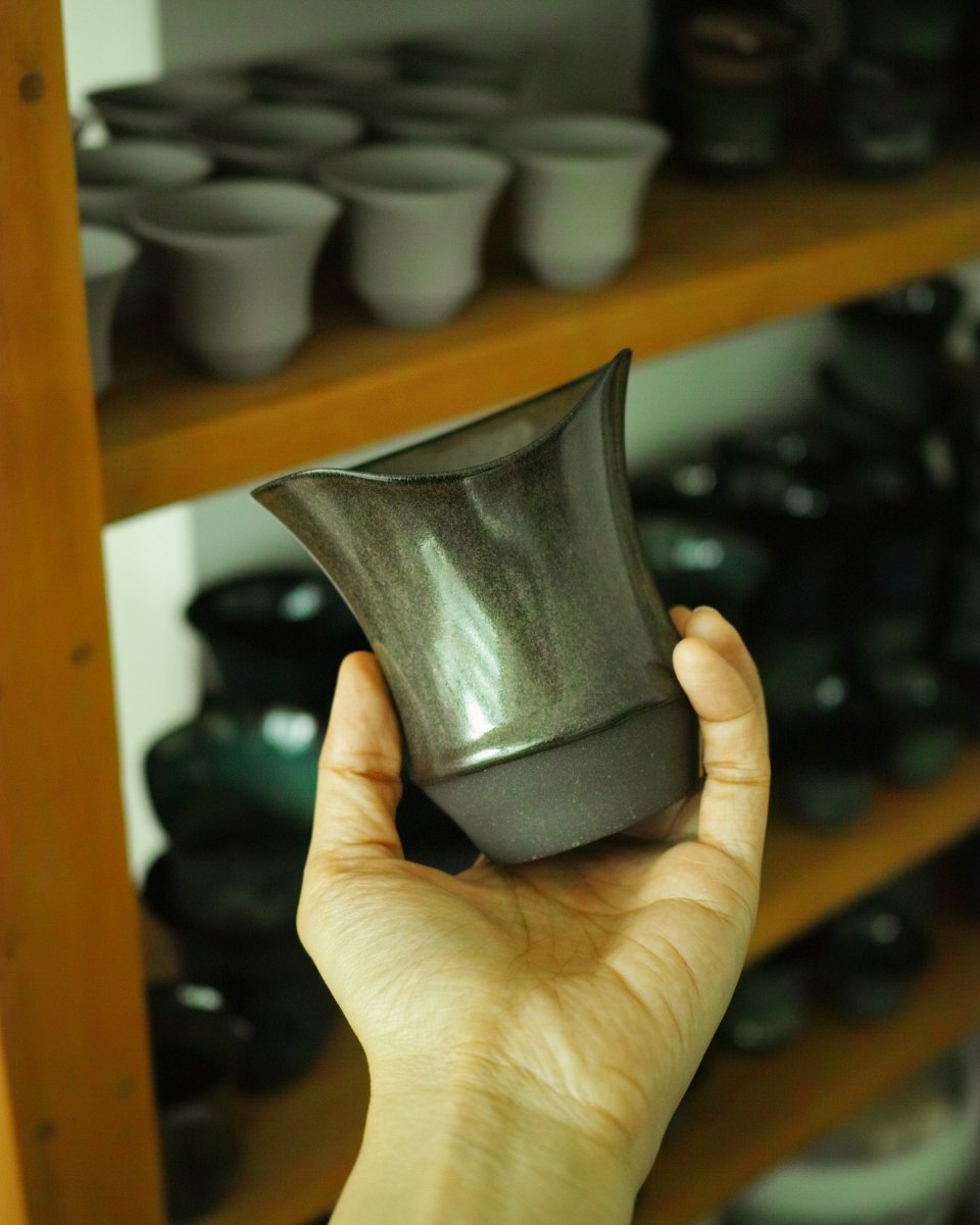 A craftsperson holding an Ember Black Gold Sharing Cup with an uneven wave-shaped rim, against a workshop rack filled with cups