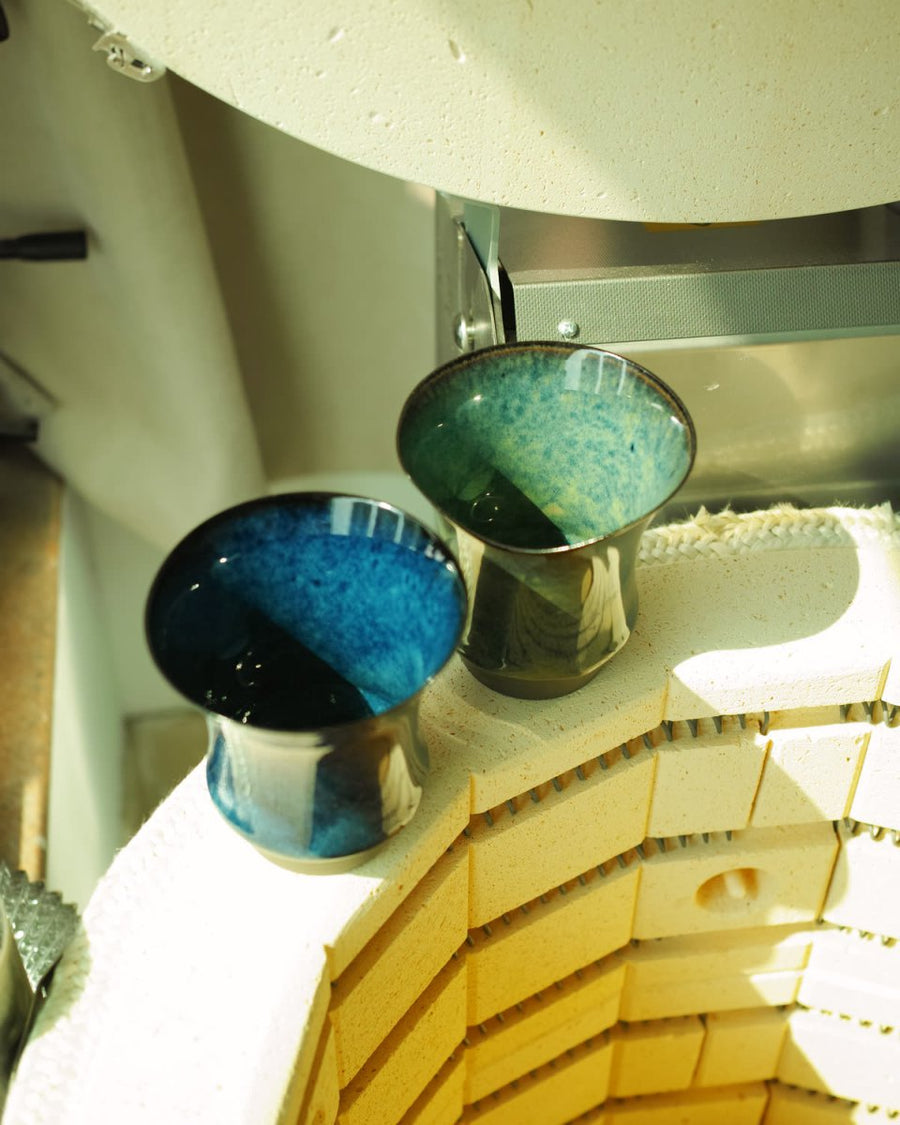 An open kiln with two freshly fired blue-green glazed cups resting at the rim
