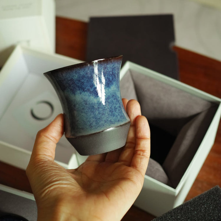 A blue-glazed cup held in front of an open white gift box with a folded grey insert
