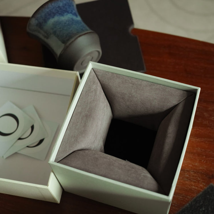 An open gift box with a folded grey insert; the cup and brand card are placed beside it