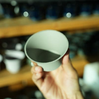 A hand lifts an unglazed cup body to inspect its inside and rim, with other cups on the workshop rack behind it