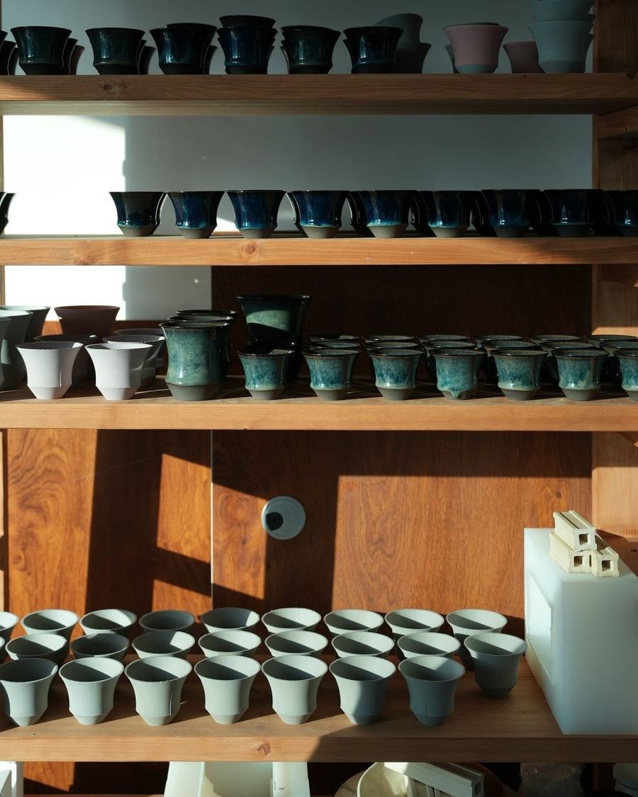 Wide view of a multi-tier workshop rack holding unglazed cup bodies and glazed fired cups together
